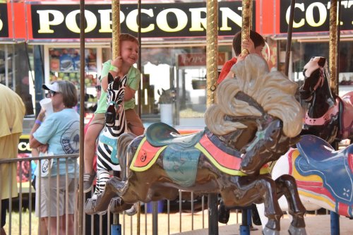 A young child joyfully rides a carousel horse at a fair, surrounded by colorful food stands and other visitors.