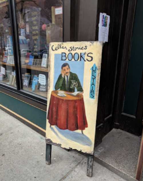 A vintage sign outside a bookstore features a man at a table with a cup, promoting "Cellar Stories Books."