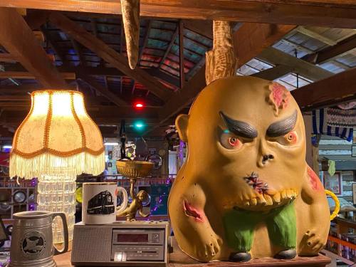 A quirky, oversized monster figurine sits among vintage decor, including a lamp and a retro radio, in a rustic setting.