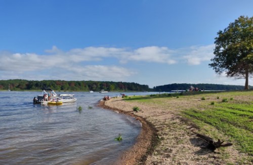A serene lakeside scene with a boat on the water, sandy shore, and green trees under a clear blue sky.