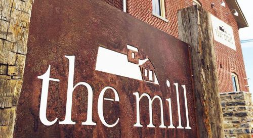 Sign for "the mill" featuring a rustic design, set against a brick building backdrop.