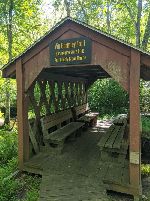 A wooden shelter at Burlingame State Park with benches, labeled "Vin Gormley Trail" and "Perry Healy Brook Bridge."