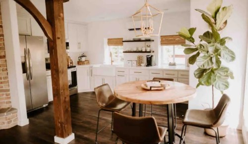 A modern kitchen with a round wooden table, four chairs, and a large plant, featuring light cabinetry and warm decor.
