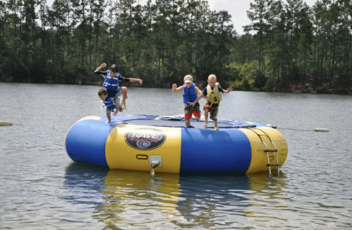 Three children jumping off a colorful inflatable water trampoline into a lake surrounded by trees.