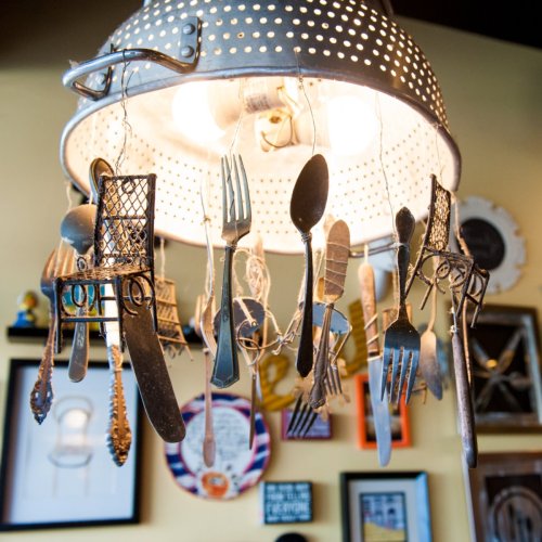 A unique light fixture made from a colander, adorned with hanging utensils like forks, spoons, and knives.
