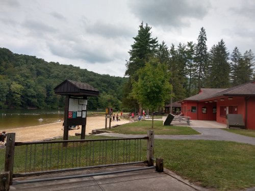 A sandy beach by a lake, with people enjoying the area and a red building nearby, surrounded by trees and cloudy skies.