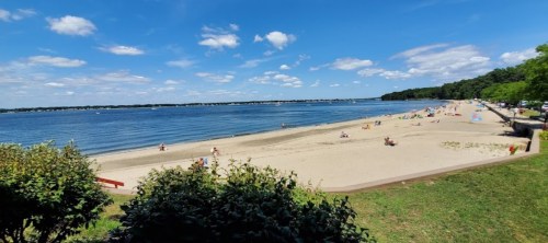 A sunny beach scene with people relaxing on the sand and calm water under a blue sky with fluffy clouds.
