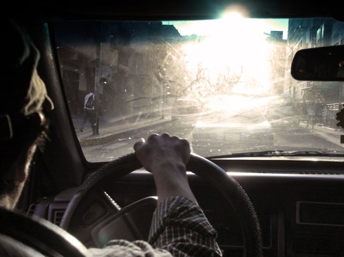 A driver's hand on the steering wheel, looking out at a sunlit, dusty street through a car windshield.