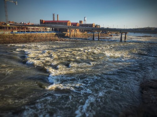 A river flows rapidly beneath a bridge, with industrial buildings in the background and a clear blue sky above.