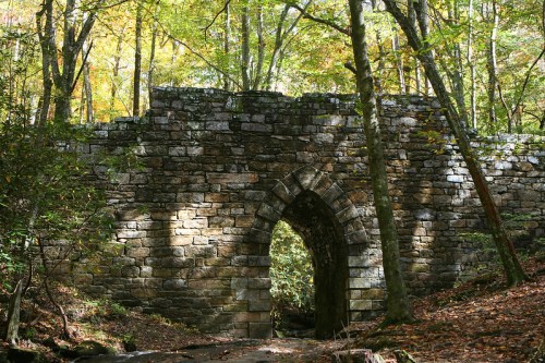Stone archway surrounded by trees, with sunlight filtering through the leaves in a serene forest setting.
