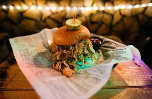 A pulled pork sandwich topped with a pickle, served on newspaper with a side of barbecue sauce.