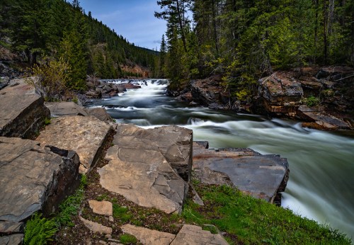 A serene river flows over rocks, surrounded by lush green trees and a mountainous landscape under a clear sky.