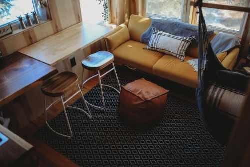 Cozy living space with a yellow couch, wooden stools, a pouf, and a patterned rug near large windows.