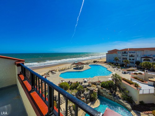 A sunny beach view featuring a pool, palm trees, and ocean waves under a clear blue sky.