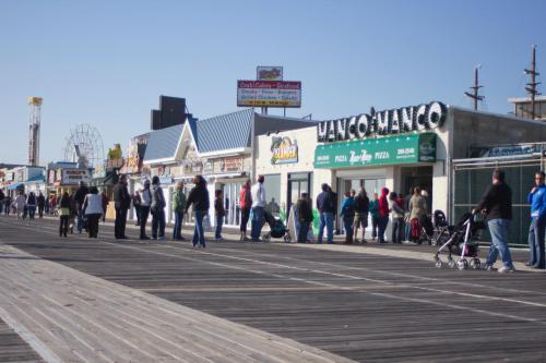 A busy boardwalk scene with people lined up outside shops, including a pizza place, under a clear blue sky.