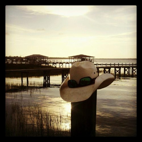 A sun hat with sunglasses rests on a post by a calm waterway, with piers and a sunset in the background.