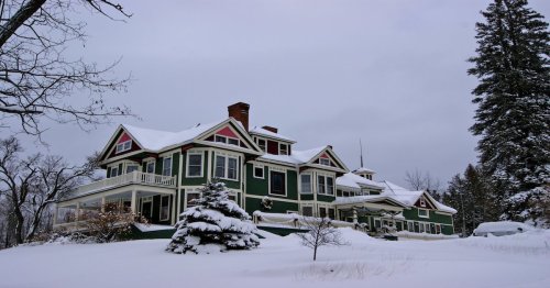A large green house with red accents surrounded by snow-covered trees and a winter landscape under a cloudy sky.
