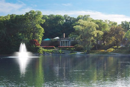 A serene lake scene with a fountain, surrounded by lush greenery and a beautiful house in the background.