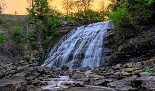 A serene waterfall cascades over rocks, surrounded by lush greenery and a warm sunset glow in the background.