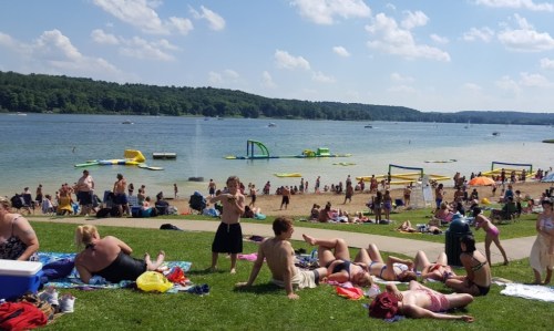A busy beach scene with people sunbathing, playing, and enjoying water activities on a sunny day by the lake.