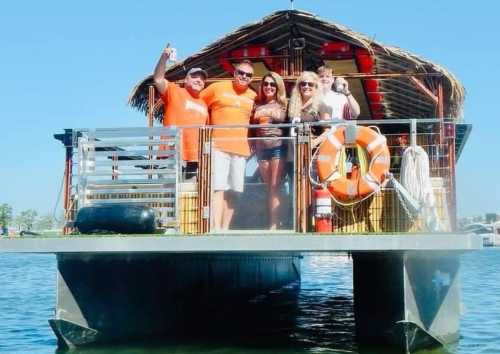 A group of five people in orange shirts smiles and poses on a boat with a thatched roof on a sunny day.