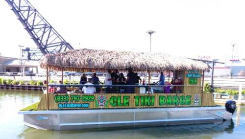 A tiki barge with a thatched roof is filled with people enjoying a sunny day on the water.