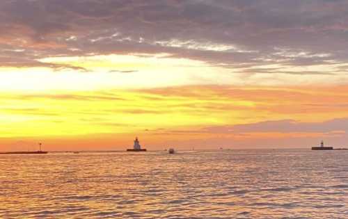 A serene sunset over water, with silhouettes of lighthouses and boats against a colorful sky.