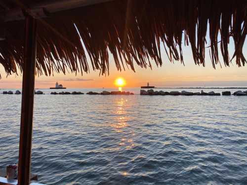 Sunset over calm waters, viewed from a thatched roof, with lighthouses in the distance and reflections on the water.