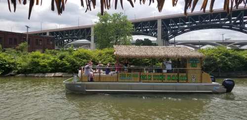 A boat with a thatched roof floats on a river, surrounded by greenery and under a bridge. People are enjoying the ride.