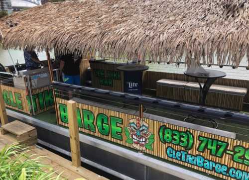 A tiki bar on a boat with a thatched roof, featuring a sign and seating area along the water.