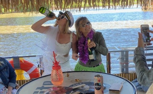 Two women in festive outfits celebrate on a boat, drinking champagne with a scenic water backdrop.