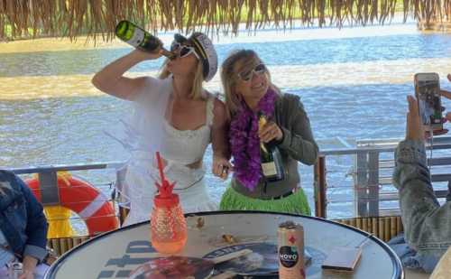 Two women in festive outfits celebrate on a boat, drinking champagne with a scenic water backdrop.