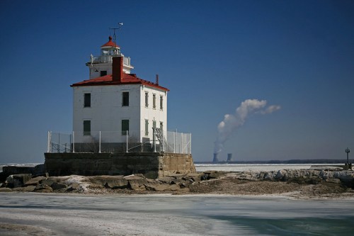 A white lighthouse with a red roof stands on a rocky shore, with smoke rising from distant power plants in the background.