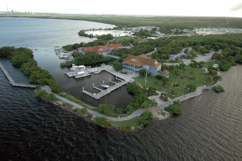 Aerial view of a waterfront property with docks, lush greenery, and a large building with red roofs.