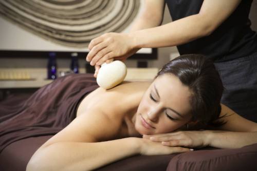 A woman receiving a massage with a stone on her shoulder, relaxing on a massage table in a serene setting.