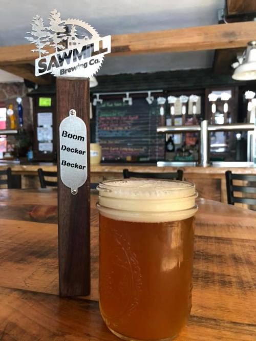 A glass of beer sits on a wooden table next to a sign for Sawmill Brewing Co. with the names "Boom," "Decker," and "Backer."