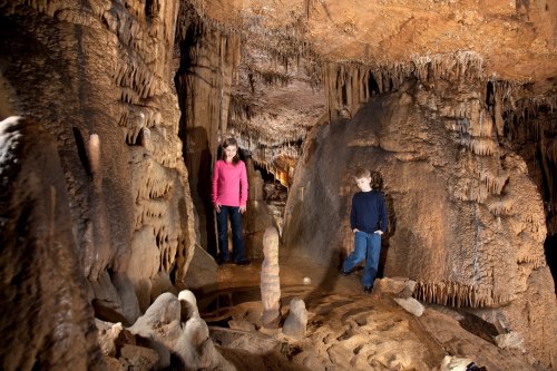 Two children explore a cave, surrounded by stalactites and stalagmites, with soft lighting illuminating the rocky formations.