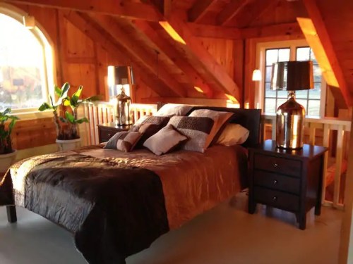 Cozy bedroom with wooden walls, a large bed, decorative pillows, and bedside tables with lamps. Natural light from windows.