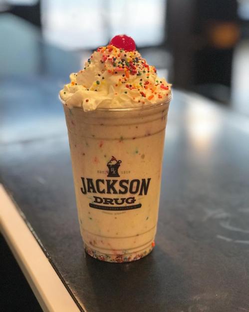 A colorful milkshake topped with whipped cream, sprinkles, and a cherry, served in a cup labeled "Jackson Drug."