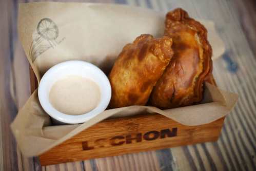 Two golden-brown empanadas in a wooden basket with a small bowl of dipping sauce.