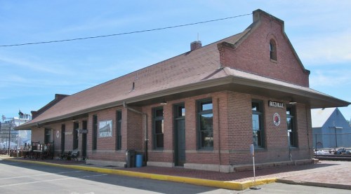 Historic brick train station with a peaked roof and large windows, set against a clear blue sky.