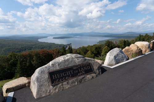 Scenic view from Height of Land, featuring a lake surrounded by lush forests and mountains under a blue sky.