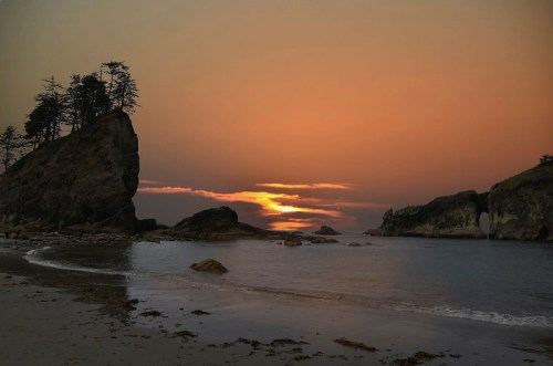 A serene beach at sunset, with rocky formations and silhouettes of trees against a colorful sky.