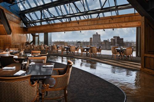 A modern restaurant with wooden decor and large windows showcasing a city skyline view. Tables are set for dining.
