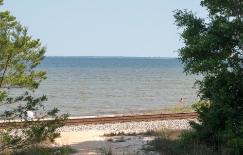 A serene view of a calm lake with a railway track in the foreground and trees framing the scene.