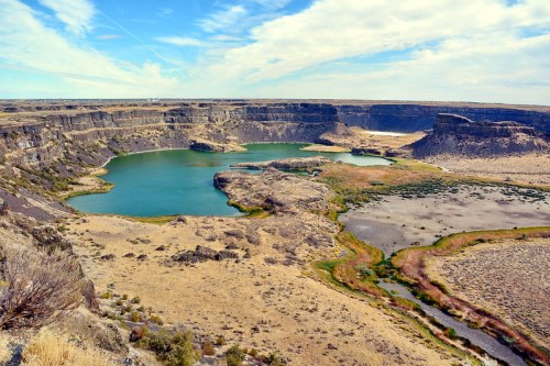 A scenic view of a canyon with a turquoise lake surrounded by rocky cliffs and dry grassland under a blue sky.