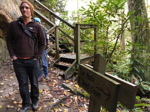 A man walks along a forest path near a wooden staircase, with a sign pointing to "The Haze" and "Sandy Cove."