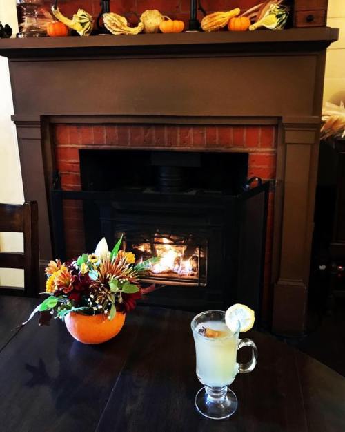 A cozy scene with a drink in front of a fireplace, decorated with autumn flowers and small pumpkins.