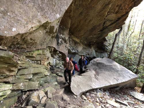 A group of hikers navigates a rocky trail beneath a large overhanging rock in a forested area.