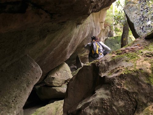 A hiker navigates through a rocky cave surrounded by lush greenery and large boulders.
